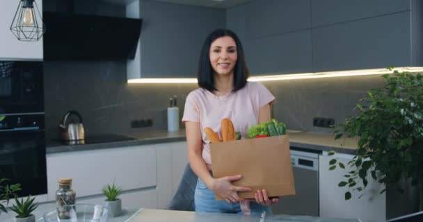 漂亮快乐的年轻女子站在现代化厨房里，一边拿着大包食物一边看着相机，一边欣赏着眼前的美景 — 图库视频影像