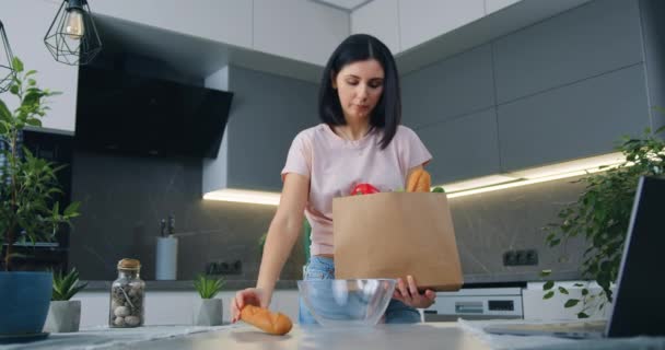 Portret dobrze wyglądającej, skoncentrowanej, 30-letniej brunetki, która wróciła ze sklepu spożywczego i rozpakowała produkty na stole kuchennym — Wideo stockowe