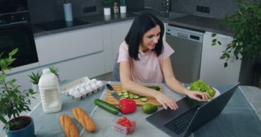 Mutfak masasında oturmuş sebze salatası hazırlarken dizüstü bilgisayar kullanarak gülümseyen genç bir esmer.
