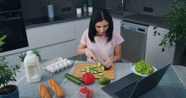 Agradável calma sorrindo jovem morena sentada à mesa na cozinha moderna e cortando pepino para salada de legumes simultaneamente escolhendo programa interessante no laptop — Vídeo de Stock