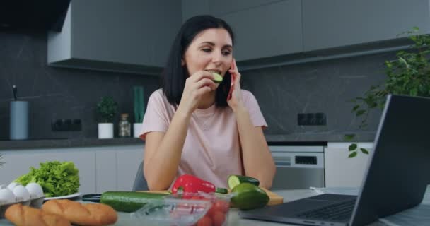 Güzel, gülümseyen, mutlu, bir dilim salatalık yiyen, telefonla konuşan ve mutfak masasında otururken dizüstü bilgisayarında ilginç bir program revize eden genç bir kadın. — Stok video