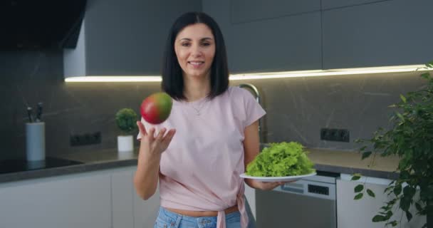 Atrakcyjny uśmiech szczęśliwa młoda brunetka stojąca w nowoczesnej kuchni i patrząc na aparat trzymając w rękach mango i liści sałaty, zdrowego odżywiania koncepcji — Wideo stockowe
