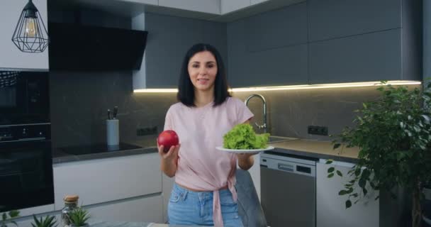 Чарівна щаслива молода темноволоса жінка позує на камеру з салатом і манго, стоячи на сучасній кухні — стокове відео