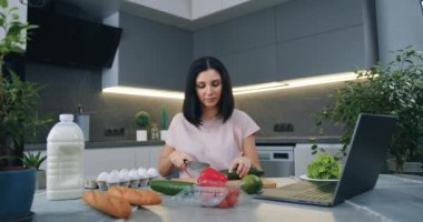 Güzel görünümlü, gülümseyen bir ev hanımı sebze salatası için salatalık kesiyor ve aynı zamanda mutfakta çevrimiçi mutfak programını izliyor.