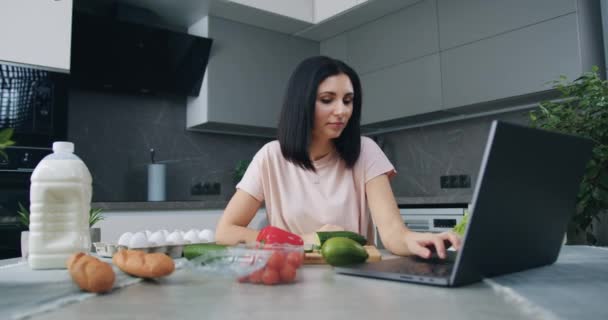 Belle jeune brune souriante et satisfaite assise à la table de cuisine et regardant le programme culinaire en ligne sur ordinateur portable pendant la préparation de la salade de légumes pour le dîner de famille — Video