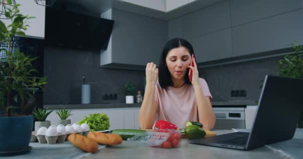 Hoş, mutlu, pozitif genç bir kadın telefonda konuşuyor ve sıcak mutfakta sebze salatası hazırlarken bir dilim salatalık yiyor. — Stok video