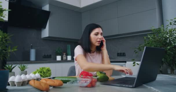 Attractive smiling confident young brunette sitting at kitchen table and working on laptop talking on phone during preparing dinner for family — Stock Video