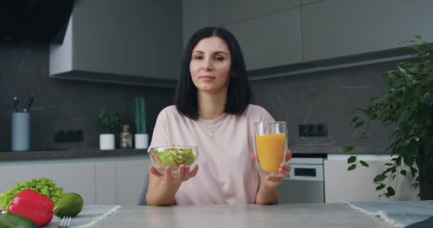 Vrij lachende tevreden jonge donkerharige vrouw zittend aan keukentafel en met een kom met salade en glas sap in haar handen — Stockvideo