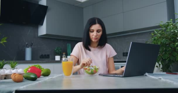 Jolie femme aux cheveux noirs concentrée de 30 ans assise à la table de cuisine, lisant des informations sur un ordinateur portable et mangeant une salade de légumes, ralenti — Video