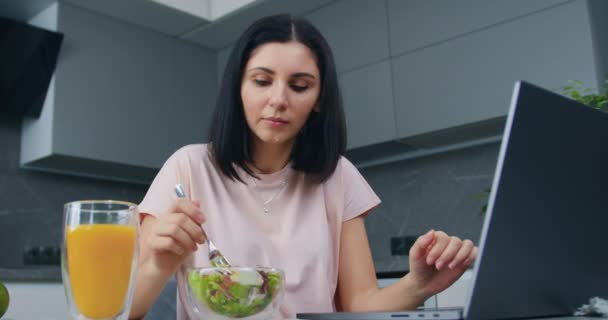Güzel, düşünceli, siyah saçlı, ev elbiseli bir iş kadını mutfak masasının yanında oturmuş laptoptan bilgi okuyor ve sebze salatası yiyor. — Stok video