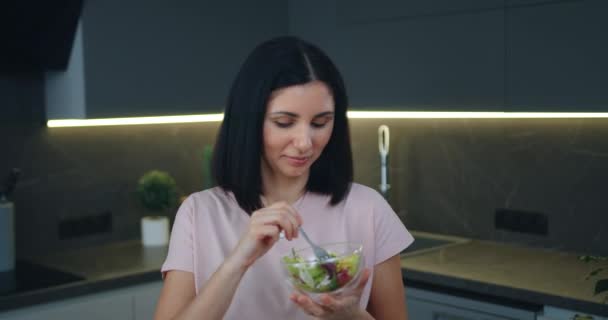 Superbe jeune femme souriante et heureuse qui mange une salade de légumes fraîchement préparée dans un bol dans une cuisine contemporaine, vue de face — Video