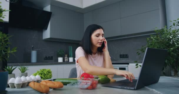 Attraente sorridente fiducioso giovane bruna seduta al tavolo della cucina e al lavoro sul computer portatile parlando al telefono durante la preparazione della cena per la famiglia — Video Stock