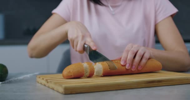 Femeia necunoscută care stă la masa de bucătărie și taie bucăți mici de baghetă proaspăt coaptă pe placa de tăiere — Videoclip de stoc