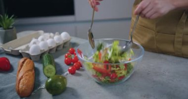 İki kaşık tutan ve şeffaf kasede sebze salatası karıştıran kadın eller mutfak masasında duruyor.