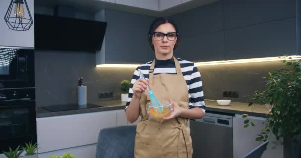 自信に満ちた若い黒髪の女性は、自分自身と彼女の家族のためにオムレツを準備泡立て器で卵を混合しながら、現代料理でカメラにポーズ — ストック動画