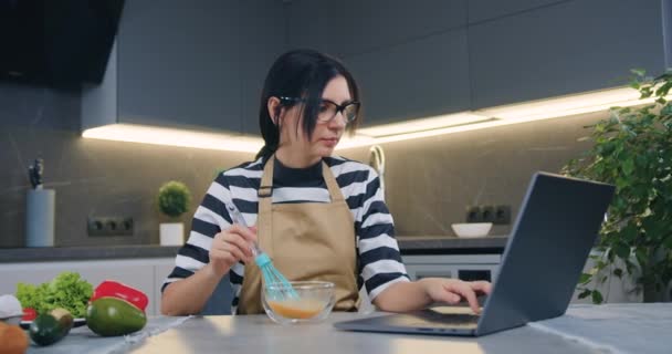 Aangename geconcentreerde 30-jarige bruneete in glazen zitten voor laptop en zoeken recept terwijl het mengen van eieren in transparante kom met garde — Stockvideo