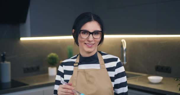 Adorable sonriente de espíritu alto mujer de 30 años en gafas mirando a la cámara mientras bate las yemas de huevo en un tazón de vidrio usando batidor — Vídeos de Stock