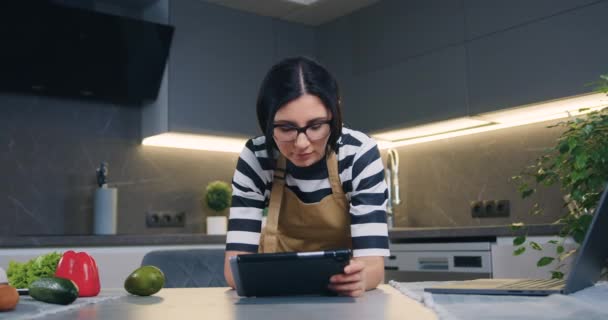 Vonzó mosolygós elégedett fiatal nő szemüvegben támaszkodik a konyhaasztalra, és böngésző alkalmazások tabletta pc, elölnézet, lassított felvétel — Stock videók