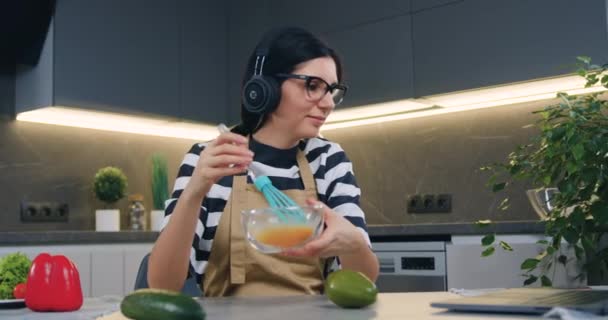 Schöne lächelnde, glückliche junge Frau mit Kopfhörern, die Musik genießt, während sie in der modernen Küche Eier in Schüssel mit Schneebesen auf dem Tisch schlägt, Frontansicht — Stockvideo