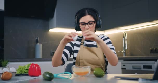 Schattig positief 30-jarige brunette slaan ei in glazen kom en zweep het met garde luisteren muziek in oortelefoons in de moderne keuken — Stockvideo
