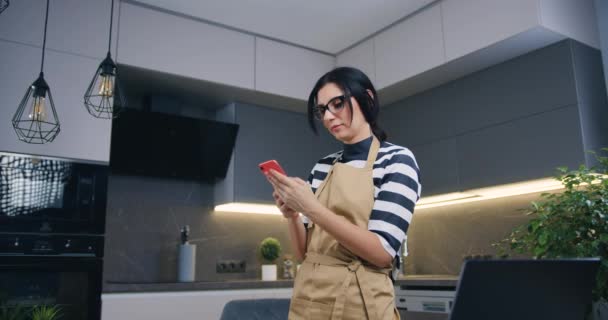 Prachtige vrolijke vrolijke jonge vrouw in glazen verheugen zich goed nieuws ze getted op de telefoon bij het staan in mooie keuken — Stockvideo