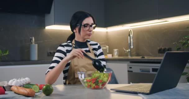 Çekici gülümseyen, koyu saçlı, kaygısız kadın cam kasede salata karıştırıyor kaşık kullanıyor ve modern mutfağın dizüstü bilgisayarında komik videolar izliyor. — Stok video