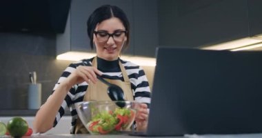 Ev yapımı hazırlık konsepti. Gözlüklü, gülümseyen, dizüstü bilgisayarda film izleyen ve kasede sebze salatasını kaşıkla karıştıran güzel bir kadın.