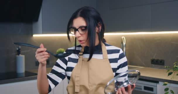 Lovely smiling satisfied 30-aged brunette in glasses preparing soup and tasting it from spoon and looking at camera with happy face — Stock Video