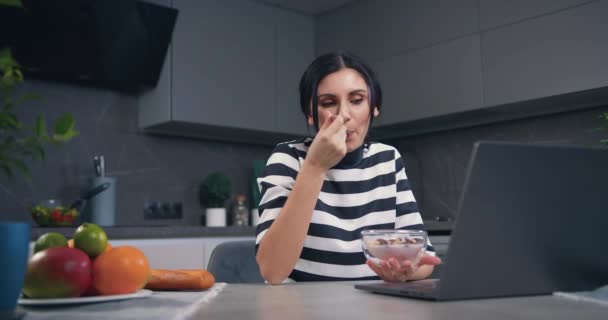 Belle jeune femme satisfaite et détendue regardant une vidéo intéressante sur ordinateur portable et mangeant des flocons de maïs avec du yaourt dans la cuisine contemporaine — Video