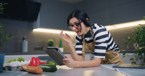 Привлекательный удивленный темноволосая молодая женщина показывает эмоциональную реакцию на хорошие новости на экране планшета, когда стоя в красивой кухне — стоковое видео