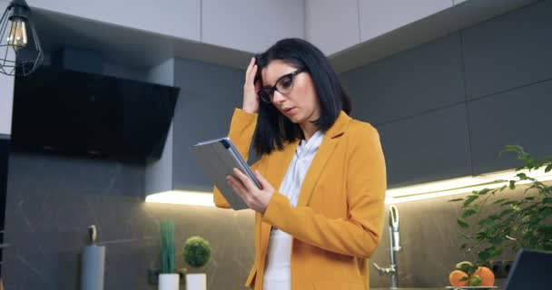 Nachdenklich sympathisch konzentrierte erwachsene schwarzhaarige Frau mit Brille und stylischem orangefarbenem Sakko steht in der gemütlichen Küche und bedient Tablet-Gerät — Stockvideo