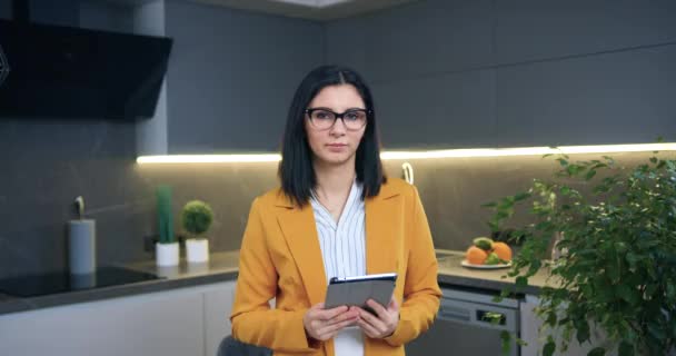 Güzel, kendine güvenen, gözlüklü ve şık ceketli başarılı iş kadını kamera önünde elinde tablet cihazla mutfak arka planında poz veriyor. — Stok video