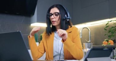 Cazibeli, kendine güvenen, kulaklıklı yetişkin bir iş kadınının modern bir dairede mutfak masasında otururken bilgisayarında video konferansı var.
