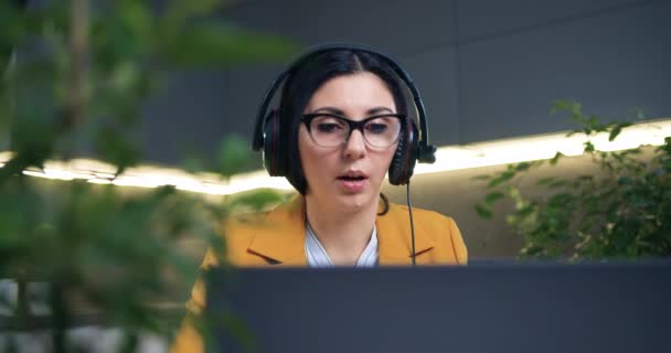 Příjemná sebevědomá úspěšná chytrá dospělá žena ve stylovém oblečení ve sluchátkách sedící před notebookem během online webináře ze speciálně vyzdobené domácí kanceláře — Stock video