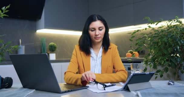 Atraente positivo confiante hábil adulto mulher de cabelos escuros em roupas elegantes colocando óculos e olhando para a câmera enquanto sentado perto do laptop na cozinha moderna — Vídeo de Stock