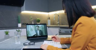 Portakal rengi ceketli güzel, attantif, deneyimli, siyah saçlı bir kadın. Çağdaş mutfakta bilgisayarın önünde oturmuş video konferans aracılığıyla iş arkadaşından açıklama yapıyor.