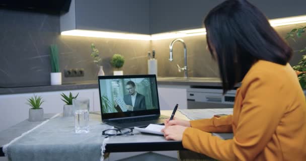 Vista posterior de la atractiva mujer de pelo oscuro exitosa attantive en chaqueta naranja que se sienta en la mesa de la cocina y tiene videoconferencia en línea con la pareja — Vídeo de stock