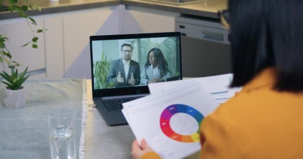 Over shoulder computer screen view of unknown dark-haired concentrated woman which sitting in front of computer during video briefing with her business partners and revisioning reports with diagramms — Stock Video