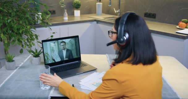 Aanzienlijke geconcentreerde geschoolde brunette in headset zitten aan keukentafel in de voorkant van de computer en het houden van videoconferentie met zakenpartner — Stockvideo