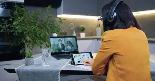 Vista posterior de una mujer morena concentrada de buen aspecto con auriculares en chaqueta naranja sentada frente a la computadora durante la reunión de video con socios internacionales y discutiendo la estrategia de negocios — Vídeo de stock