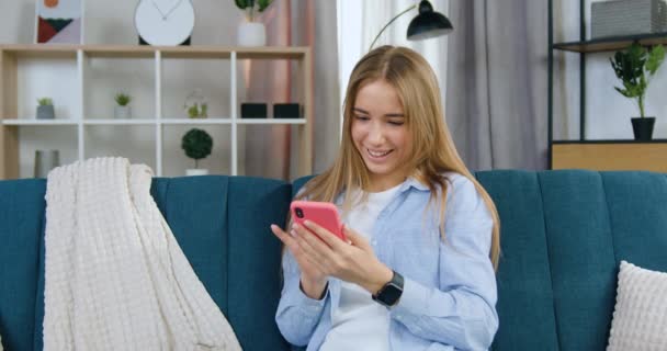 Linda feliz sorrindo despreocupado 20-aged loirinho em roupas casuais sentado no sofá macio na sala de estar e escrevendo mensagem no telefone para sua amiga, close-up — Vídeo de Stock