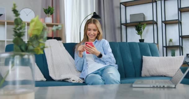 Charmigt leende glad ung flicka med långt hår i avslappnade kläder sitter på soffan i modernt vardagsrum och revidera roliga videor på mobilen — Stockvideo