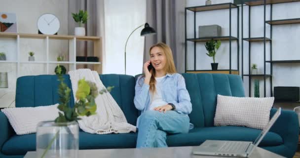 Attraktivt leende glad ung ljus hår flicka i casual kläder sitter på soffan hemma och har positiva telefonsamtal, framifrån — Stockvideo