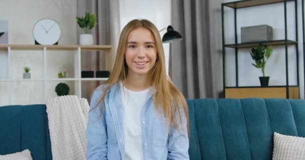 Imádnivaló mosolygós boldog fiatal szőke hajú lány kék ingben ül a kanapén a kamera előtt, miközben pózol a kortárs lakás, elölnézet — Stock videók