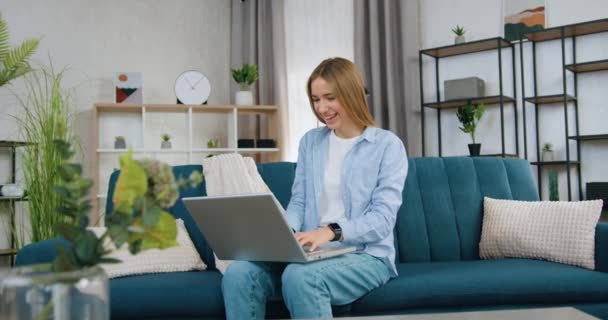 Attraktiv positiv nöjd ung blond hår flicka sitter på mjuk soffa i samtida vardagsrum och arbetar på datorn, slow motion — Stockvideo