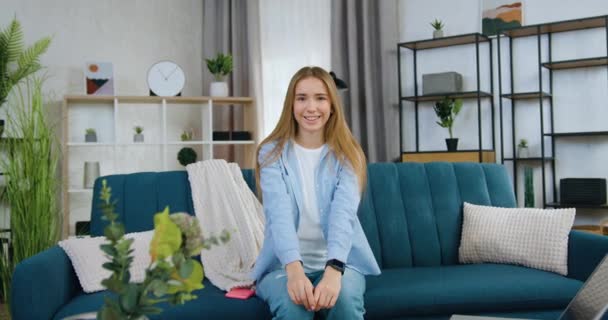 Boa aparência sorridente feliz menina elegante com cabelos longos e lisos sentado no sofá confortável na sala de estar e olhando para a câmera com rosto satisfeito — Vídeo de Stock