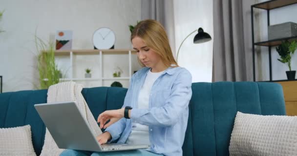 Uroczy w dobrym nastroju inteligentny młody jasnowłosy student kobieta w ubraniach casual siedzi na kanapie w domu i pracuje na laptopie, pisząc domowy uniwersytet — Wideo stockowe