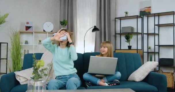 Hermoso positivo moderno dos diferentes edades niñas pasar ocio conjunto en jugar juegos virtuales con el ordenador portátil y gafas de realidad aumentada, vista frontal — Vídeo de stock