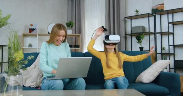 Charmoso alegre luz cabelos 20-aged menina escolher entretenimento virtual para sua irmã mais nova, que está sentado perto dela no sofá macio em óculos de realidade aumentada — Vídeo de Stock