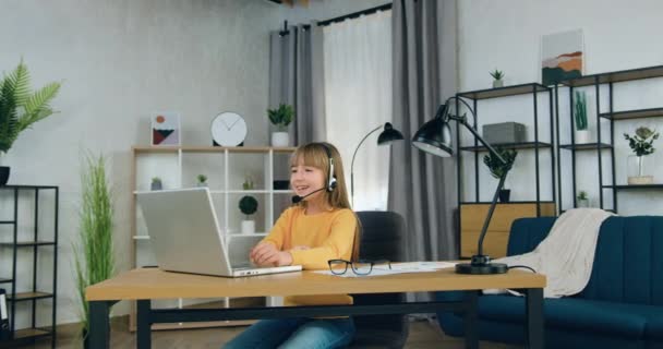 Boa aparência positiva confiante júnior menina em fones de ouvido sentado na frente do laptop e conversando com o professor durante reunião de vídeo de casa, conceito de educação escolar remota — Vídeo de Stock